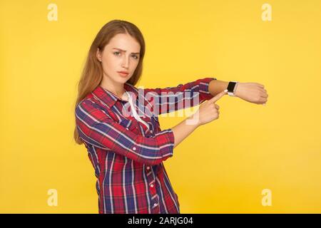 Regardez l'heure ! Portrait d'une jeune fille de gingembre sérieuse dans une chemise à carreaux pointant vers la montre sur son poignet et regardant décontent, se préoccupant de la fin de l'heure. Banque D'Images