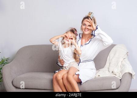 Joyeuse maman heureuse et fille avec des bigoudis dans les cheveux regardant l'appareil-photo à travers les doigts dans les jumelles geste et souriant se amuser ensemble sur le canapé, gr Banque D'Images