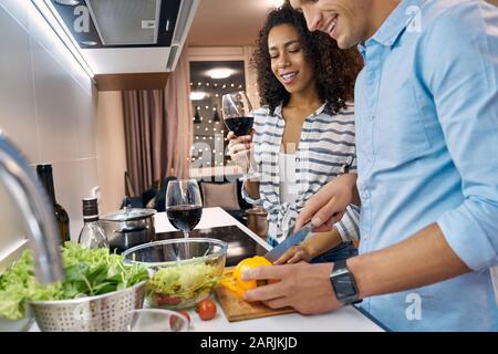 Date Romantique. Jeune couple multiethnique debout à la cuisine cuisine dîner femme boire du vin tandis que mari couper des légumes gros plan rire Banque D'Images