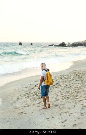 Guy touriste avec un sac à dos jaune marche le long de la plage de sable près de l'océan. Banque D'Images