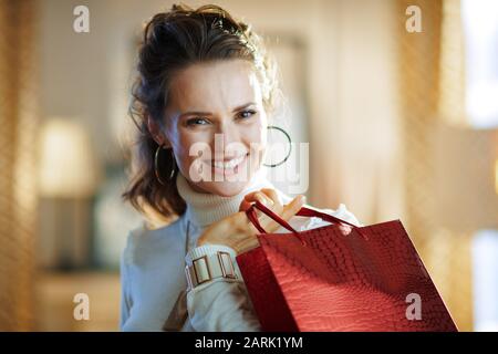 Portrait d'une femme souriante et élégante de 40 ans en pull blanc et jupe avec sac de shopping rouge dans la maison moderne en hiver ensoleillé. Banque D'Images