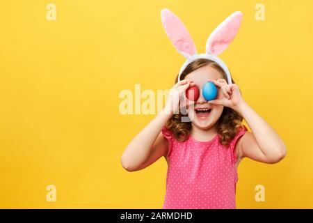 Un enfant joyeux et joyeux aux oreilles d'un lapin tient deux œufs de Pâques dans les yeux. Portrait d'une petite fille sur un fond jaune dans le studio. Banque D'Images