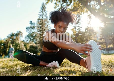 Portrait d'une jeune femme afro-américaine déterminée assise sur l'herbe verte le matin lumière du soleil étirant ses jambes regardant l'appareil photo Banque D'Images