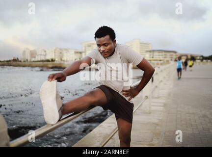 Déterminé homme se courbant pour toucher les orteils tout en mettant la jambe sur la rampe tout en s'exerçant près de la mer le matin Banque D'Images
