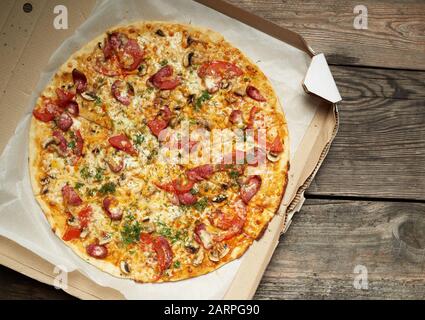 Ronde au four pizza avec saucisses fumées, champignons, tomates, fromage et de l'aneth dans une boîte en carton sur une table en bois, de l'alimentation est coupée en portions, t Banque D'Images