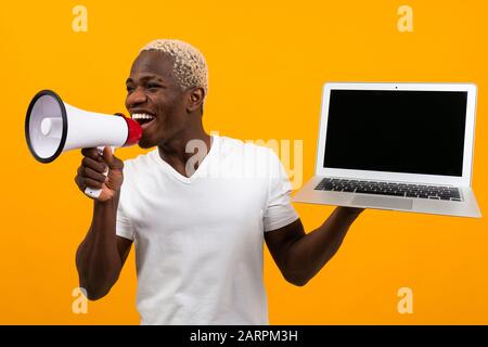 un homme noir africain avec des cheveux blancs parle dans un mégaphone tenant un ordinateur portable avec une maquette pour la publicité sur un fond de studio jaune. Banque D'Images