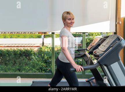 Une femme d'âge moyen qui s'exerce sur un tapis roulant et regarde l'appareil photo. Salle de gym avec vue sur la nature sur le parc vert le jour ensoleillé. Image couleur avec espace de copie. Santé Banque D'Images