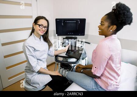 Heureuse femme souriante médecin montrant l'échographie de bébé sur tablette numérique à sa patiente à la clinique. Bonne femme africaine enceinte ayant Banque D'Images