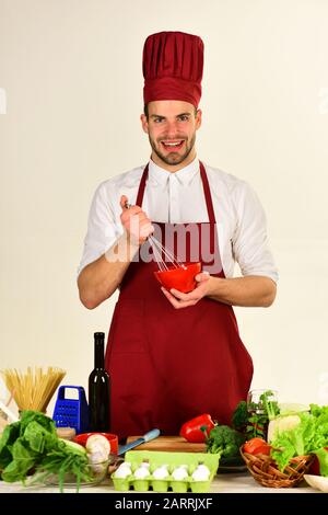 L'homme dans le chapeau de cuisinier et le tablier tient des ustensiles de cuisine. Cuisiner travaille dans la cuisine près de la table avec des légumes et des outils. Le chef au visage heureux tient le bol et fouetter sur fond blanc. Cuisine et concept de cuisine Banque D'Images