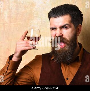 Guy avec verre de cognac. Macho avec brandy de boissons au visage flambant sale ou whisky. Concept de service et de restauration. L'homme avec barbe et moustache détient une boisson alcoolisée sur fond de mur beige. Banque D'Images