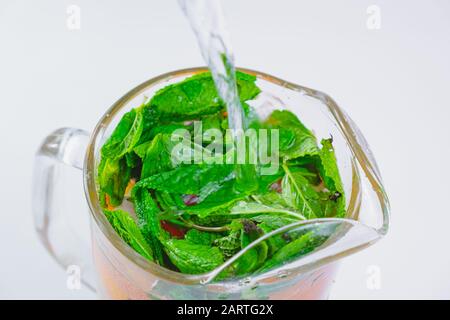 Verser de l'eau sur des feuilles de menthe poivrée fraîche et des tranches de pamplemousse dans une vue rapprochée en verre. Préparer de l'eau tonique ou de la limonade, saine boisson fraîche con Banque D'Images