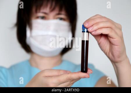 Tube à essai dans la main femelle près, femme dans le masque médical tenant un flacon avec du liquide rouge. Concept de l'échantillon sanguin, de la vaccination, de la recherche sur le coronavirus Banque D'Images