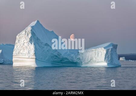Énorme iceberg flottant dans un fjord au coucher du soleil avec lune au milieu, fjord, Scoresby Sund, Kangertittitaq, Groenland,Danemark Banque D'Images