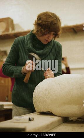 1970, sculpteur historique et féminin à l'intérieur d'un atelier sur un banc de travail sur une pièce en pierre incurvée avec marteau et petit burin, Angleterre, Royaume-Uni. La sculpture fait partie des arts visuels, avec une sculpture durable un processus où une œuvre est faite de sculpture et de modélisation d'une gamme de matériaux tels que la pierre. Banque D'Images