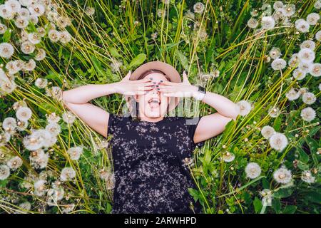 Jeune belle fille dans les lunettes de soleil se trouve dans le champ de pissenlit et fermer les yeux avec plaisir. Vue de dessus. Banque D'Images