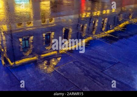 Réflexion floue d'un bâtiment lumineux dans une flaque. Pluie dans la ville la nuit. Arrière-plan coloré et abstrait. Mise au point sélective Banque D'Images