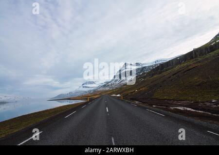 Route entourée par la rivière et les collines couvertes dans le Neige et herbe sous un ciel nuageux en Islande Banque D'Images