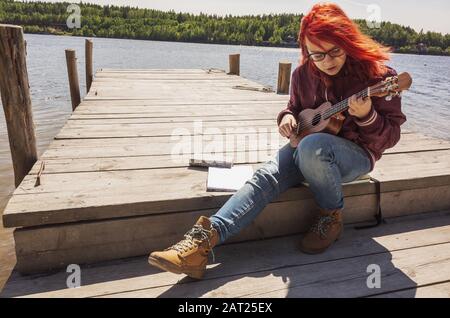 Une jeune fille caucasienne dans des verres avec des cheveux rouge vif joue ukulele sur une jetée en bois Banque D'Images