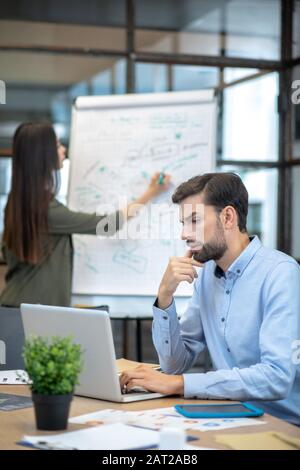 Homme barbu dans une chemise bleue assise à la table et travaillant sur un ordinateur portable Banque D'Images