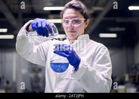 Gros plan jeune femme de laboratoire dans des verres et un manteau blanc avec ballon avec liquide bleu dans ses mains vérifie la qualité sur fond défocté Banque D'Images