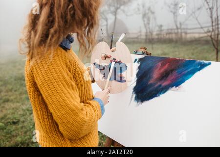 Gros plan photo de la jeune artiste féminine travaillant sur la peinture à l'extérieur. Elle contient des peintures à l'huile, des brosses artistiques, de la toile et de la palette. Elle mélange des couleurs Banque D'Images