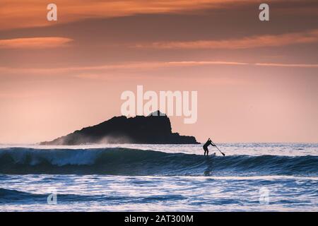 Une paddle-boarder paddling près de Goose Rock silhouetted par le feu de fin de soirée à Fistral à Newquay à Cornwall Banque D'Images