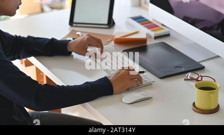 Photo latérale du jeune homme créatif en tapant sur le clavier. Y compris tasse à café, souris, clavier, guide de couleur, écouteurs pour tablette, ordinateur portable et crayon Banque D'Images