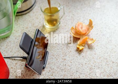 recharge sans fil pour smartphone sur le socle de chargement à côté de la tasse à thé en verre sur le plan d'examen de la cuisine. Banque D'Images