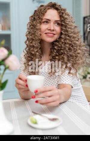 Bonne fille blonde avec des boucles assis dans un café avec une tasse de thé. Portrait intérieur de la jolie dame qui se trouve à la table Banque D'Images