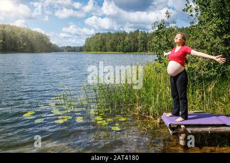 La jeune femme enceinte pond sur un ponton en bois sur un lac forestier pendant une chaude journée ensoleillée se répandant ses mains en profitant de la nature - sain grossesse vie Banque D'Images