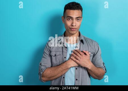 Beau homme mulatto regardant la caméra tenant ses mains sur sa poitrine debout contre un fond bleu - image Banque D'Images