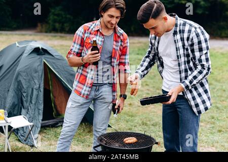 Deux gars cuisinant une saucisse sur le barbecue dans le camping. - Image Banque D'Images