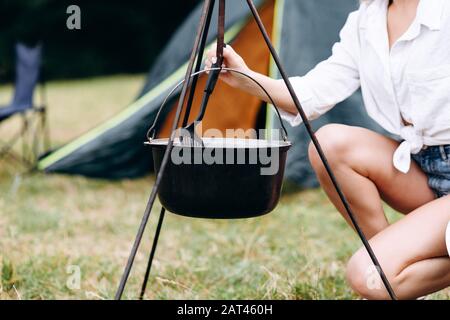 Jeune femme cuisine dans le camping assis à côté du feu de joie - recadrage de l'image Banque D'Images