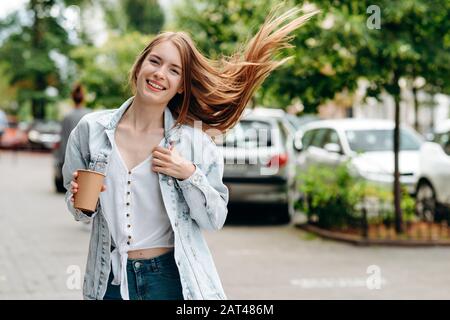 Fille souriante à cheveux rouges debout à l'extérieur tenant un café en papier verre - image Banque D'Images