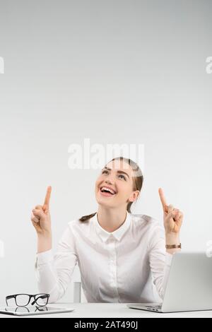 Une femme heureuse est assise à côté d'un ordinateur portable et rire semble pointer les doigts vers le haut sur ses deux mains. - image verticale Banque D'Images