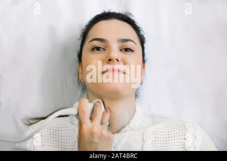 Vue rapprochée du visage de la jeune femme caucasienne au cours de l'examen échographique d'une thyroïde posée sur le canapé dans le cabinet médical Banque D'Images