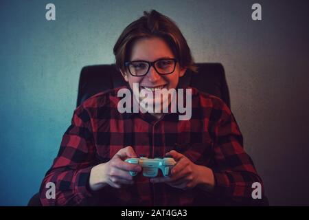 Joyeux adolescent jouant à des jeux vidéo assis dans son fauteuil. Un adolescent excité portant des lunettes, tenant la console du joystick sourire à la caméra, essayez t Banque D'Images