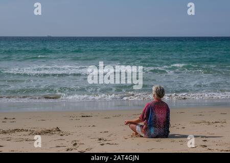 Femme senior avec des cheveux gris et une robe blanche se relaxant sur les cheveux de plage médite sur la plage Banque D'Images