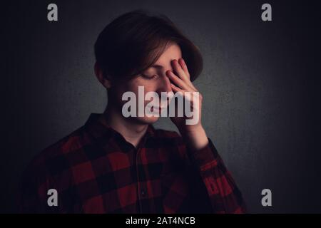 Un adolescent affligé s'agite dans sa tête, pointant vers le front, les yeux ont fermé la douleur, isolé sur fond gris mur sombre de la salle. Maux de tête fe Banque D'Images