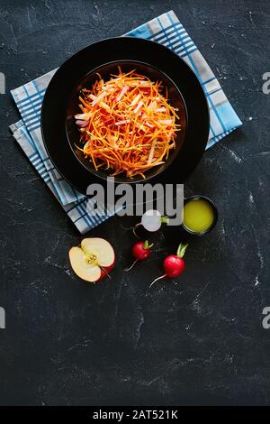 Salade fraîche de radis et de pomme aux carottes saupoudrée de graines de quinoa et de zeste de citron dans un bol noir sur une table en bois avec ingrédients, vue verticale Banque D'Images