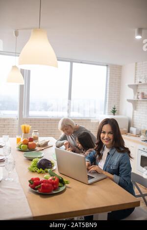 Maman avec ordinateur portable, grand-mère et petite-fille le matin dans la cuisine. Banque D'Images