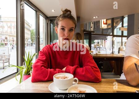 Jeune femme assise dans un café avec un grand gobelet cappuccino sur la table. Prise de vue moyenne. Regarder l'appareil photo. Banque D'Images