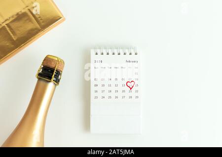 Carte de Saint-Valentin, bouteille de champagne dorée, chocolat dans un wrapper or et calendrier de février avec une date entourée sur une surface blanche. Vue de dessus. Banque D'Images