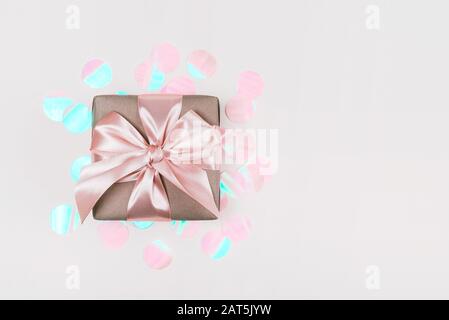 Boîte cadeau enveloppée dans du papier d'artisanat avec ruban rose parfait sur fond rustique rose pâle avec des confettis holographiques. Banque D'Images