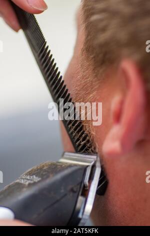 Coiffeur rasage de la barbe et des whiskers avec tondeuse à cheveux ou tondeuse à cheveux dans un salon de coiffure professionnel ou un barbershop , vu de derrière le client, non reconnaissable. Banque D'Images