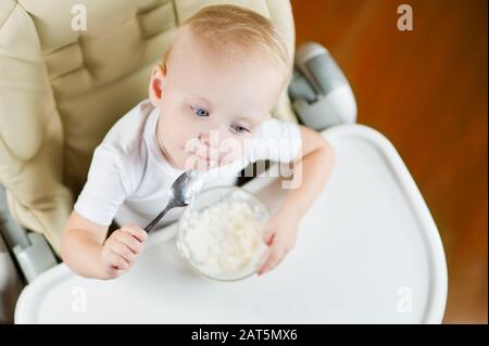 Une petite fille mange du porridge avec une cuillère, assis sur une chaise d'alimentation avec une table blanche et pense à quelque chose. Vue de dessus Banque D'Images