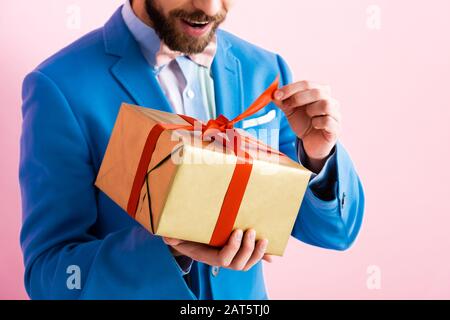 vue rognée de l'homme excité dans le costume touchant ruban sur la boîte cadeau isolée sur rose Banque D'Images