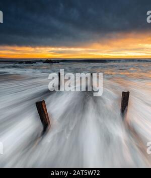 Avec l'eau blanche se déplaçant rapidement après 4 pylônes, l'image est équilibrée par le ciel lourd plein de pluie au-dessus de la bande vibrante de lumière de coucher d'or. Banque D'Images