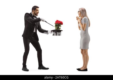 Photo pleine longueur d'un magicien faisant un tour magique avec des fleurs et un chapeau devant une jeune femme surprise isolée sur fond blanc Banque D'Images