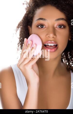 surprise fille africaine américaine avec des bretelles utilisant la brosse faciale nettoyante en silicone, isolée sur blanc Banque D'Images